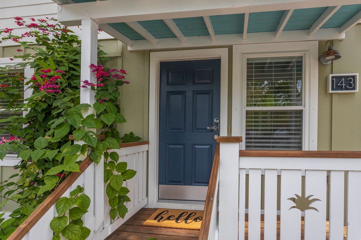 The entryway features a dark blue door framed by lush greenery and vibrant pink flowers. A welcome mat lies on the wooden steps, while a light blue overhang provides shade. The house number is displayed on the wall beside the door.