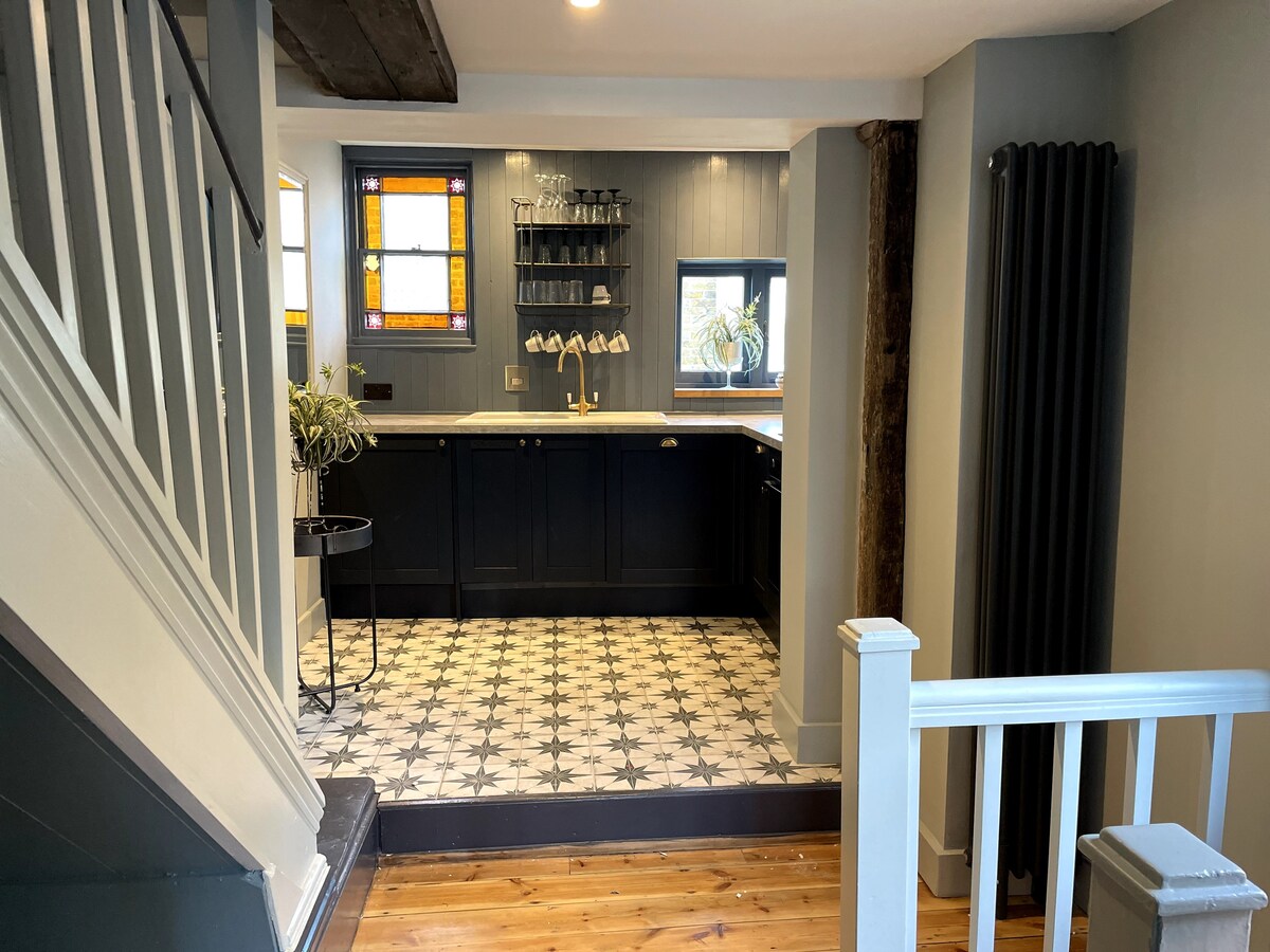 The open plan kitchen area features a warm tiled floor with a star pattern and modern black cabinetry. Natural light enters through a decorative stained glass window, illuminating the space. A modern radiator and rustic wood accents add character to the inviting kitchen environment.