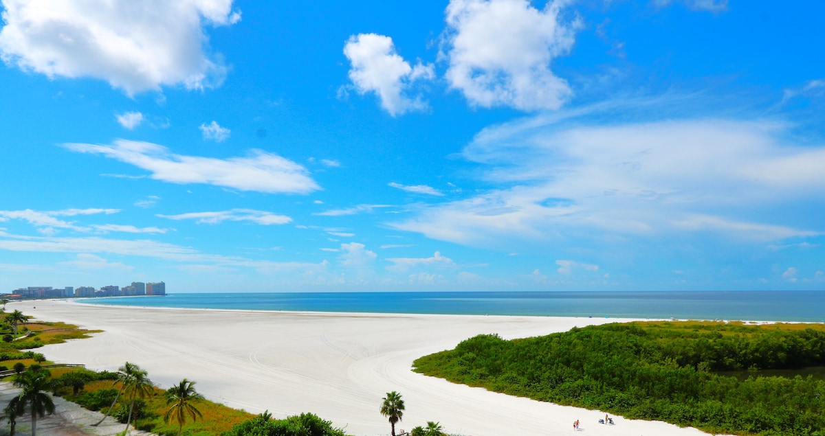 A wide view of Marco Island's Crescent Beach is presented, featuring soft white sand meeting the calm blue waters. Green vegetation lines the shore, while distant buildings can be seen against a bright sky with scattered clouds.