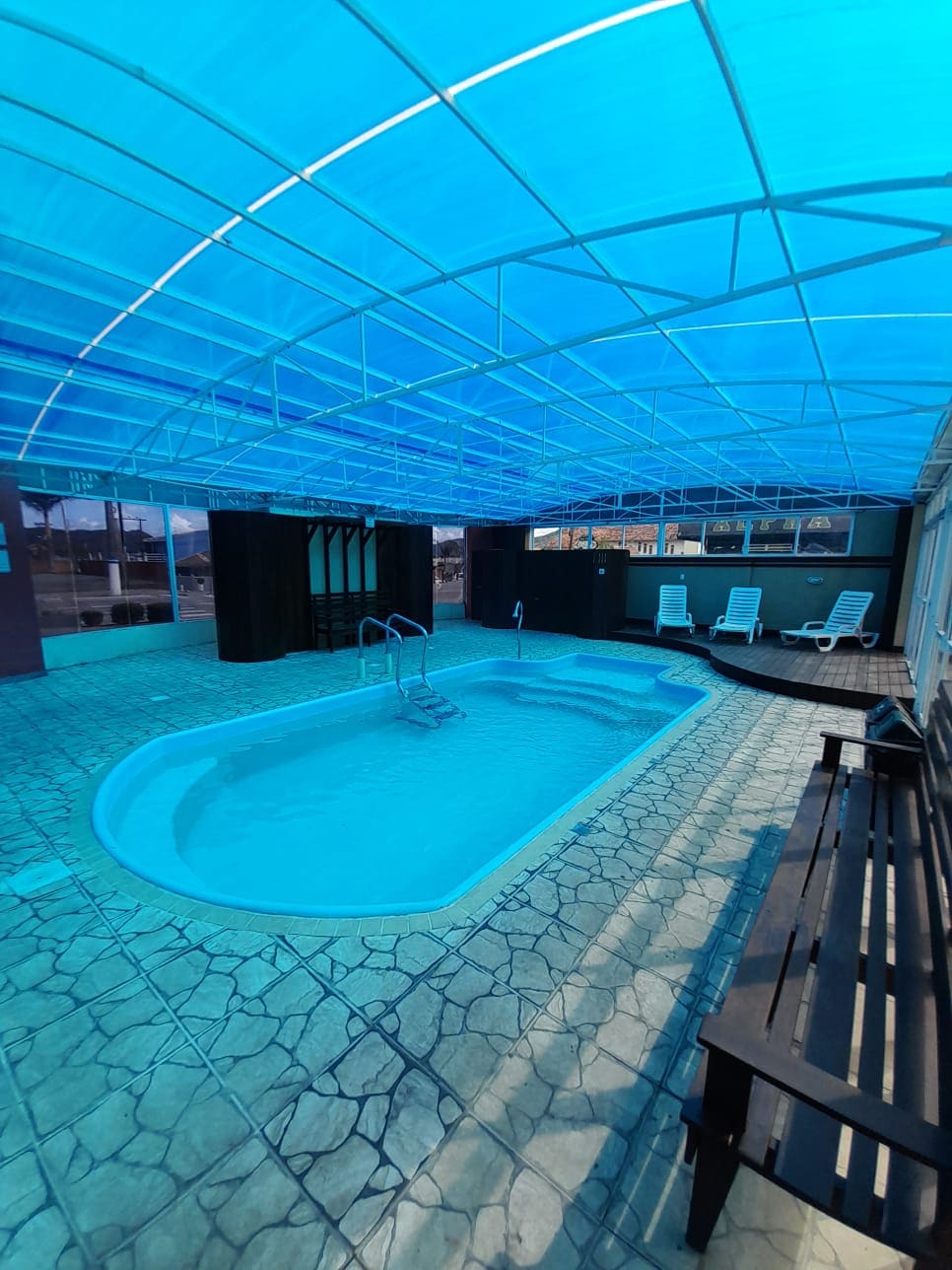 An indoor pool area is featured, showcasing a clear blue water swimming pool surrounded by a textured stone floor. Sunlight filters through a transparent ceiling, illuminating the space. Lounge chairs are positioned nearby, enhancing the relaxation environment.