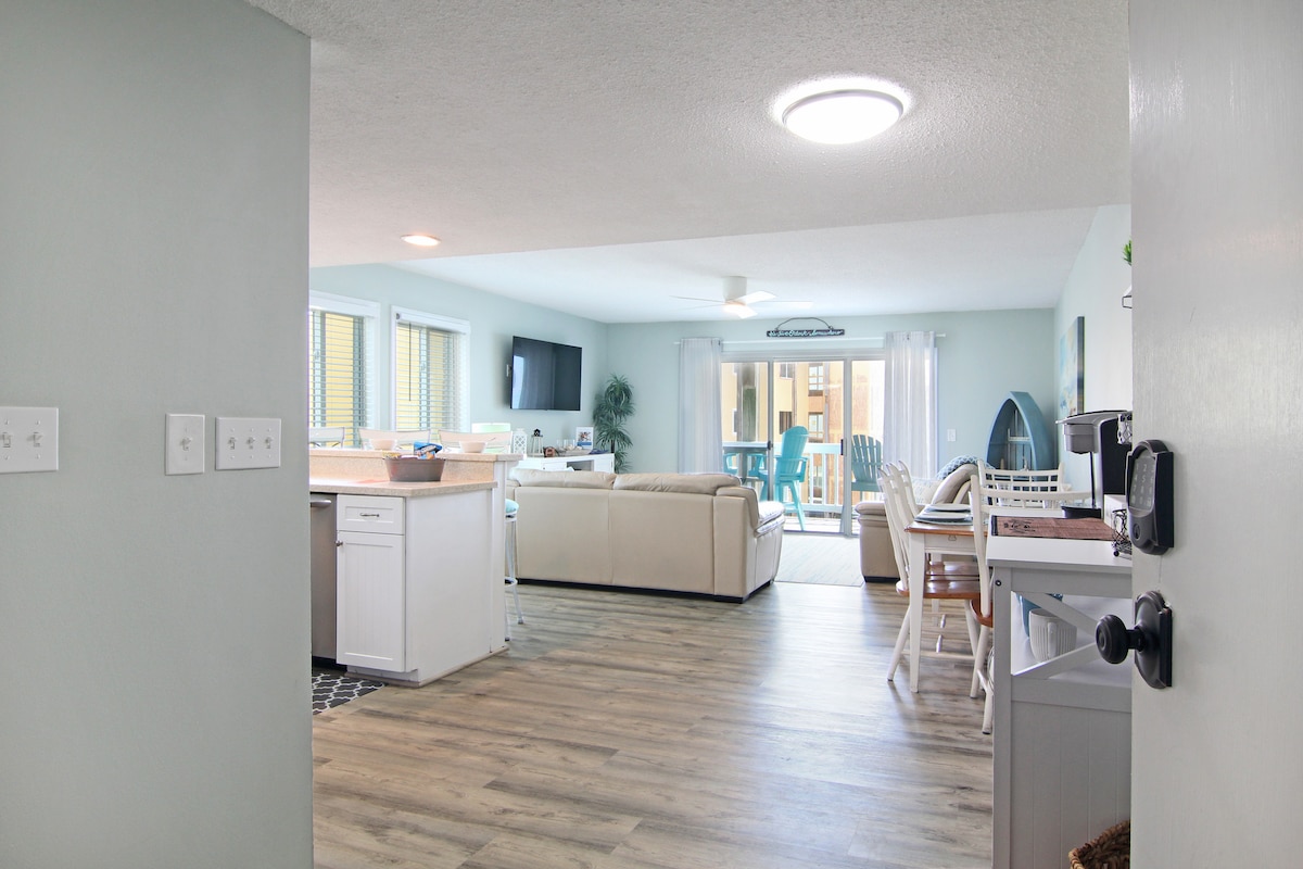 A spacious living and dining area is visible, featuring light wood flooring and soft pastel walls. A well-equipped kitchen with white cabinets opens into the comfortable seating area. Large sliding glass doors lead to a covered porch, allowing natural light to fill the space.
