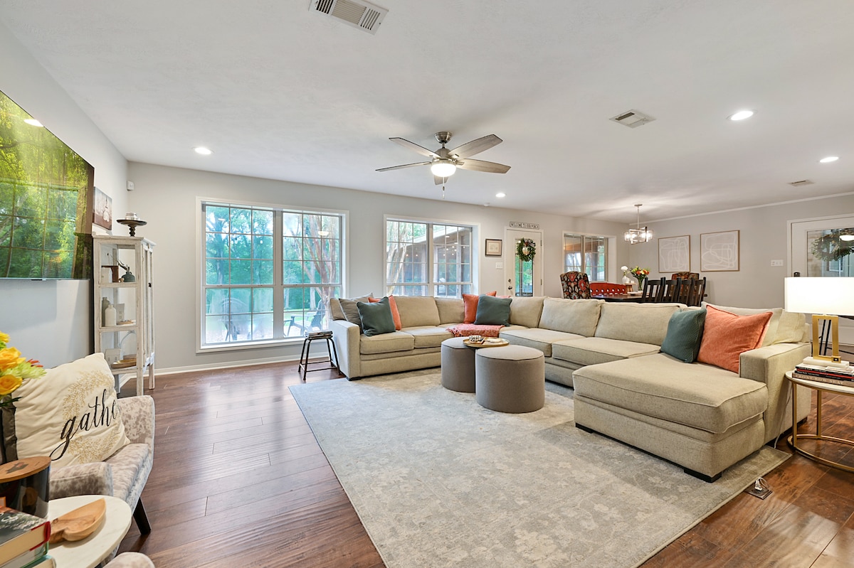 A spacious living area features an oversized sectional couch arranged around a circular coffee table. Large windows bring in natural light, revealing views of greenery outside. A ceiling fan and modern lighting fixtures enhance the room's comfort, while decorative pillows add a touch of color.