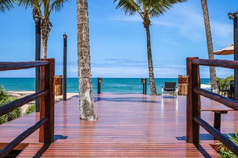 A wooden deck leads toward the ocean, framed by palm trees swaying gently in the breeze. Wooden railings line the deck, and a sun lounger is positioned to enjoy the coastal views. A clear blue sky complements the serene beach atmosphere.