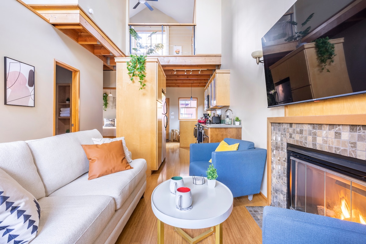 An open concept living area showcases a cozy cream sofa with colorful accent pillows. A round coffee table is placed in front, accompanied by two blue armchairs. Light streams in through windows, highlighting the warm wood accents and a wall-mounted TV above a stone fireplace.