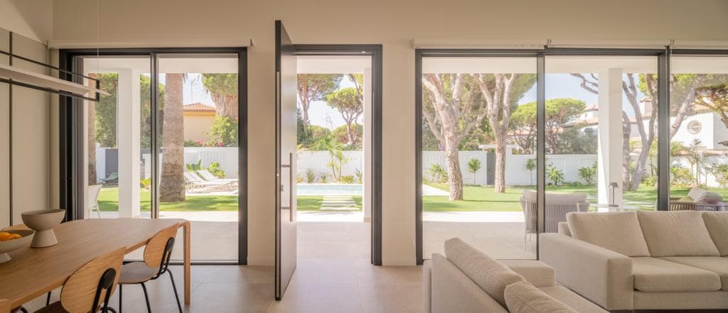 A spacious living area is showcased, featuring large sliding glass doors that open to the outer garden and pool area. Natural light fills the interior, where a modern dining table is set alongside a comfortable seating area, overlooking the landscaped outdoors.
