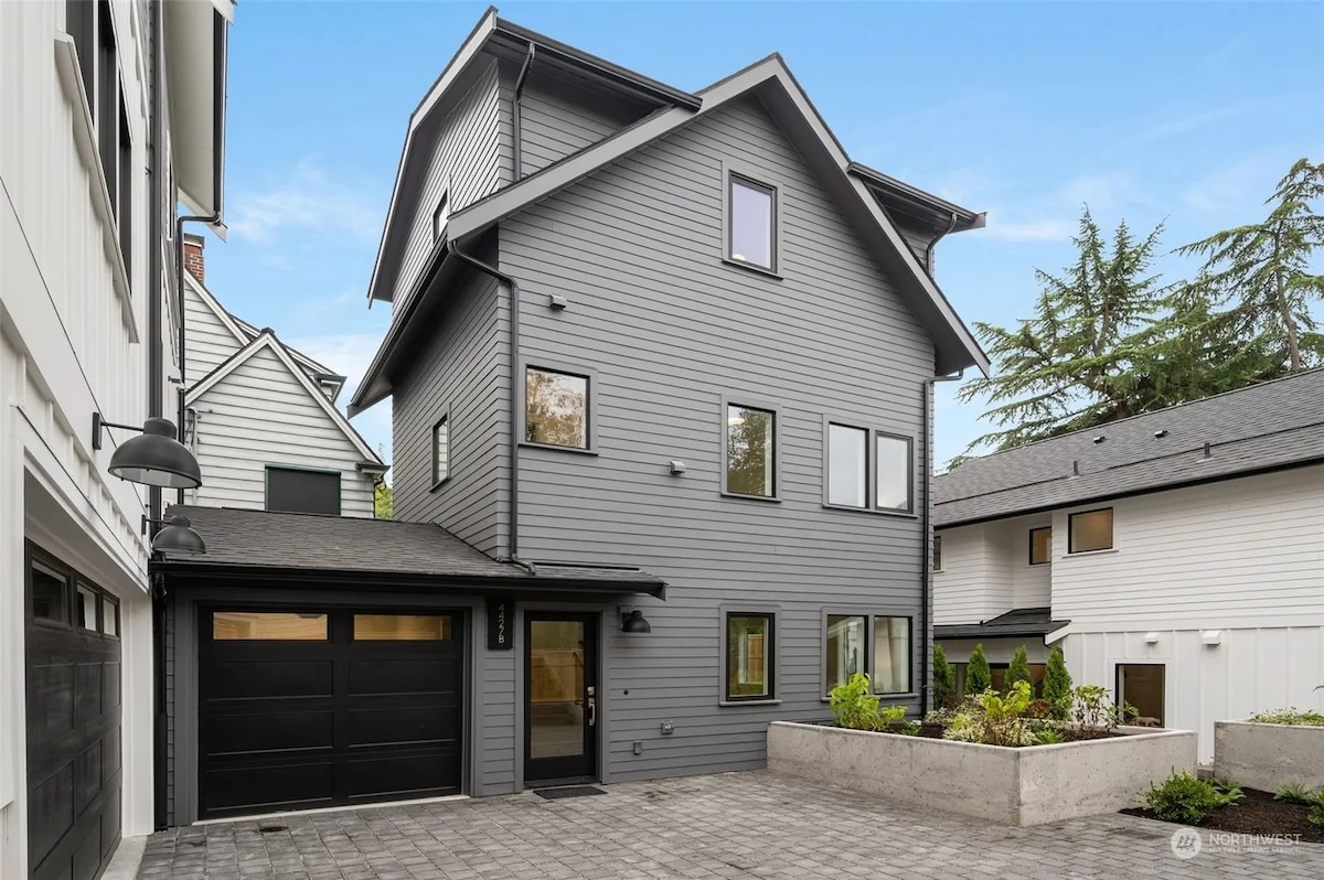 A contemporary gray house is showcased, featuring a sleek black garage door and large windows. Outdoor landscaping includes a small garden area with stone borders, while the surrounding space is paved with square tiles, enhancing the modern aesthetic.