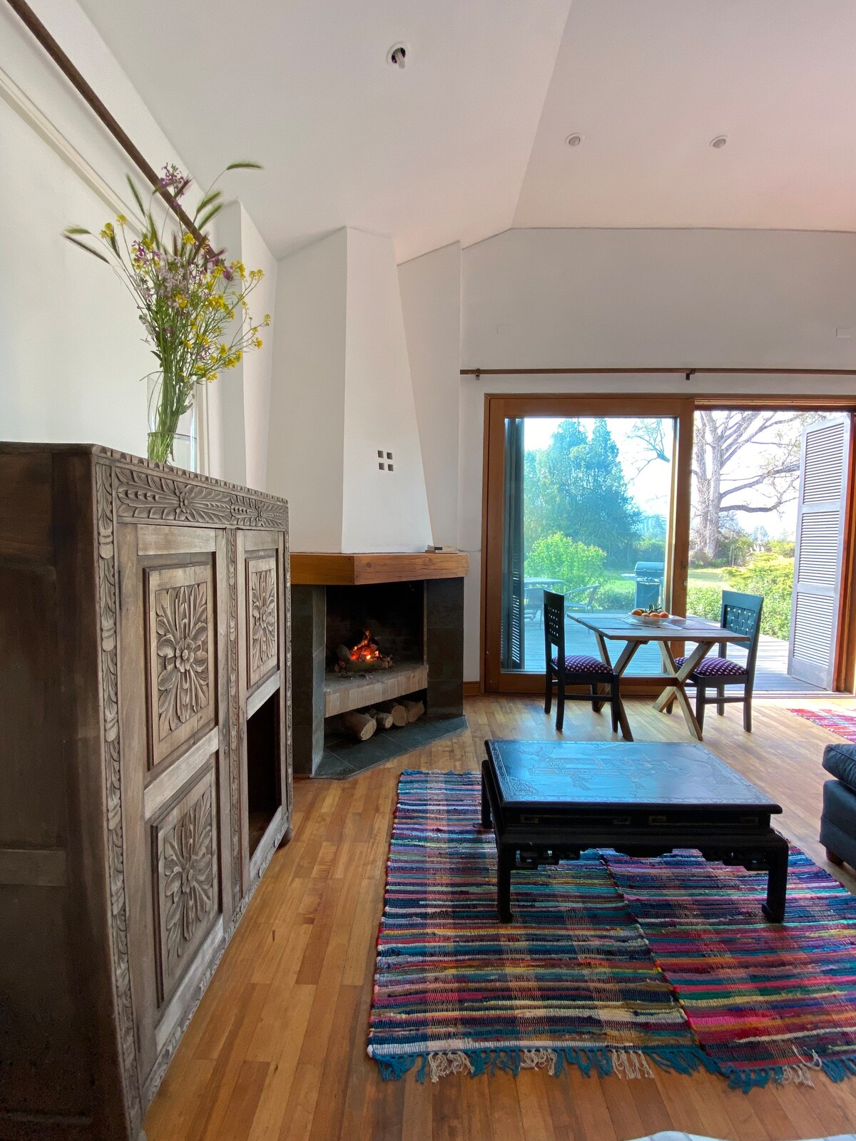A cozy living area is highlighted by a wood-burning fireplace and a decorative cabinet. Natural light enters through large sliding doors, providing a view of the garden outside. A wooden coffee table and dining set complement the colorful striped rug on the hardwood floor.