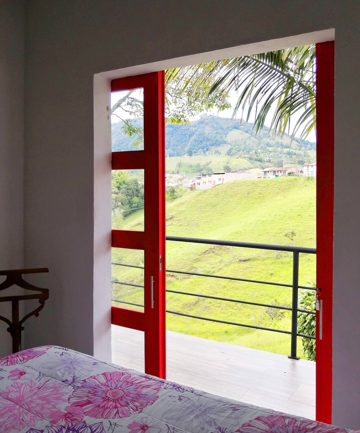 A bright red door frames a scenic view of lush green hills and distant structures. The door opens onto a balcony, enhancing the connection to the natural surroundings. A flower-patterned comforter is visible in the foreground.