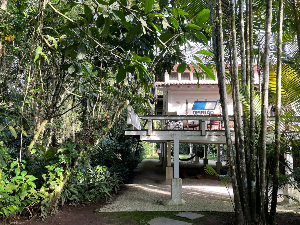 A wooden house on stilts is nestled among lush greenery, with trees and tropical plants surrounding it. The structure features a deck area, promoting a connection with nature. Gravel pathways lead to the house, emphasizing the tranquil, natural setting.
