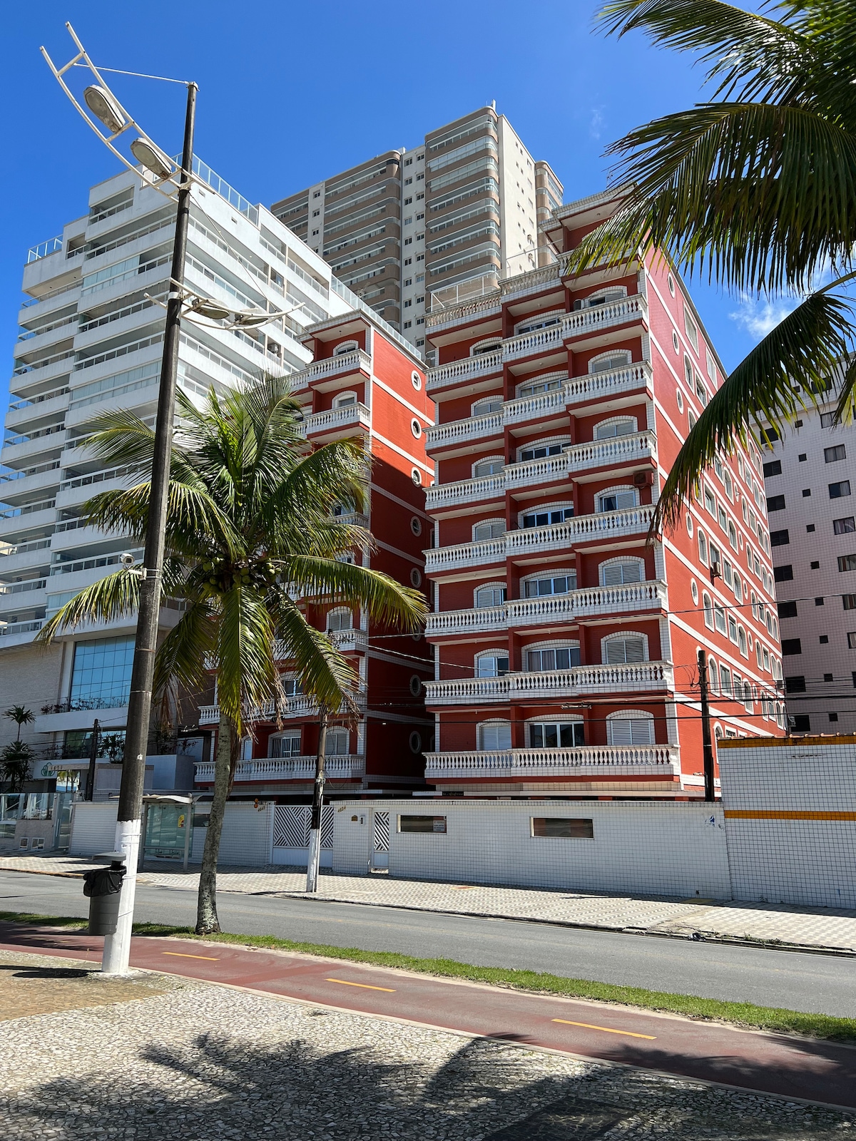 A multi-story red building is visible, showcasing distinct architectural features with balconies on each floor. Tall palm trees are present in the foreground, contributing to a tropical atmosphere. Nearby, additional modern structures create a contrasting skyline against a clear blue sky.