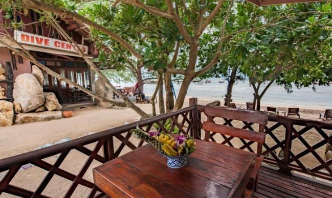 An outdoor seating area is highlighted by a wooden table adorned with a bowl of tropical fruits. Surrounding greenery provides shade, creating a peaceful retreat. In the background, the sandy beach and water are visible, suggesting a serene coastal atmosphere.