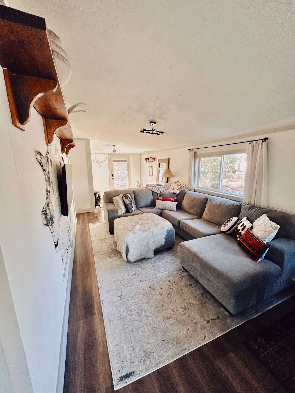 A spacious living area is presented with a luxury sectional positioned on a soft rug. Natural light filters through two large windows, illuminating decorative accents on the walls. A wooden shelf displays small decorative items, contributing to a cozy and inviting environment.