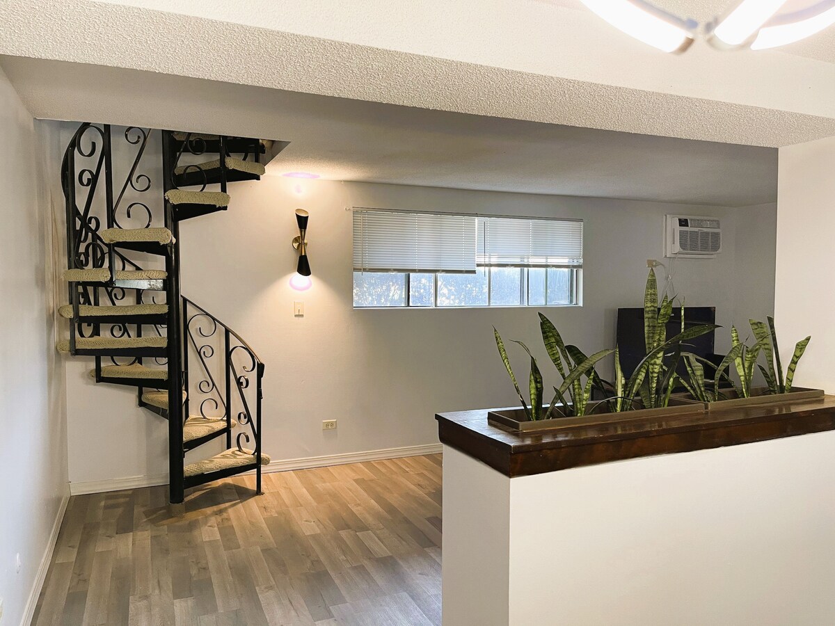 A spacious living area is highlighted by a decorative spiral staircase leading to the upper level. Natural light filters through the window, illuminating the room. Green plants are positioned along the low wooden railing, adding a touch of nature to the decor.