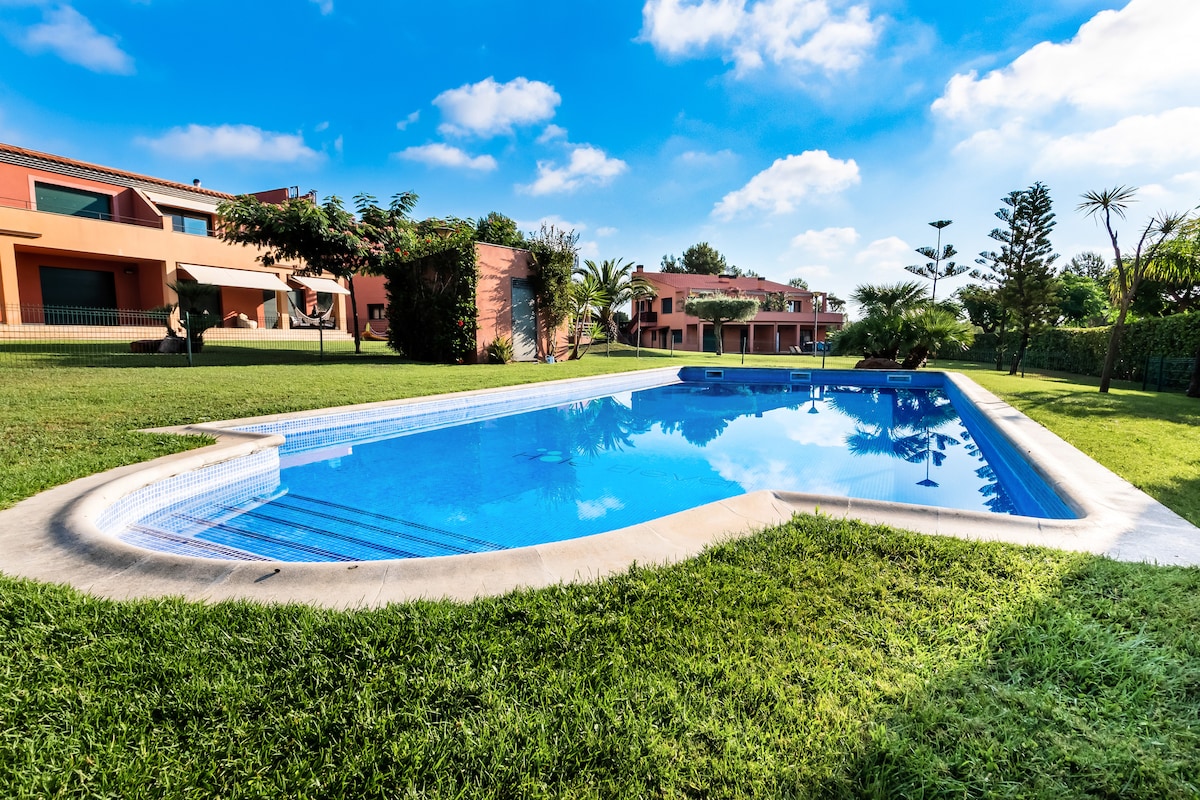 A clear, inviting swimming pool is surrounded by a well-maintained green lawn and palm trees. Soft sunlight reflects off the water's surface, while additional residential buildings are visible in the background, providing a serene environment for relaxation.
