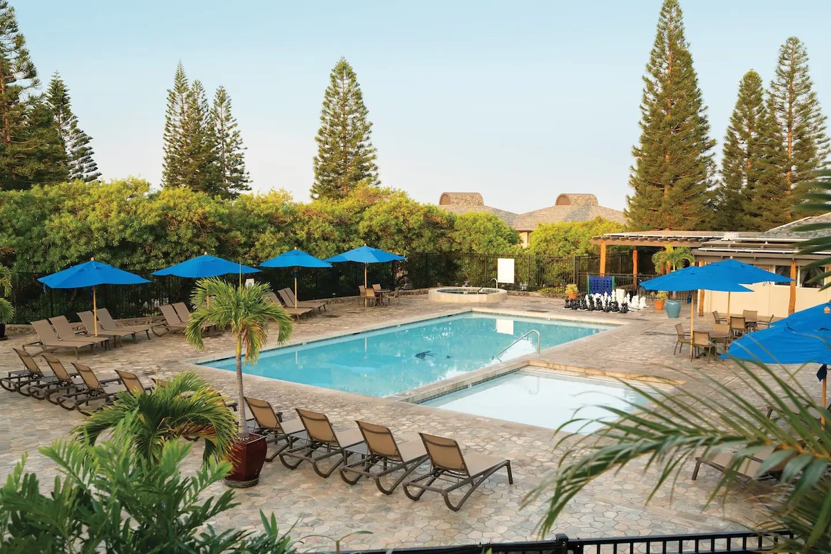 The outdoor swimming pool area is surrounded by lush greenery and features a tiled patio with numerous lounge chairs. Bright blue umbrellas provide shade over some chairs. A hot tub is positioned near the pool, enhancing the relaxing atmosphere.