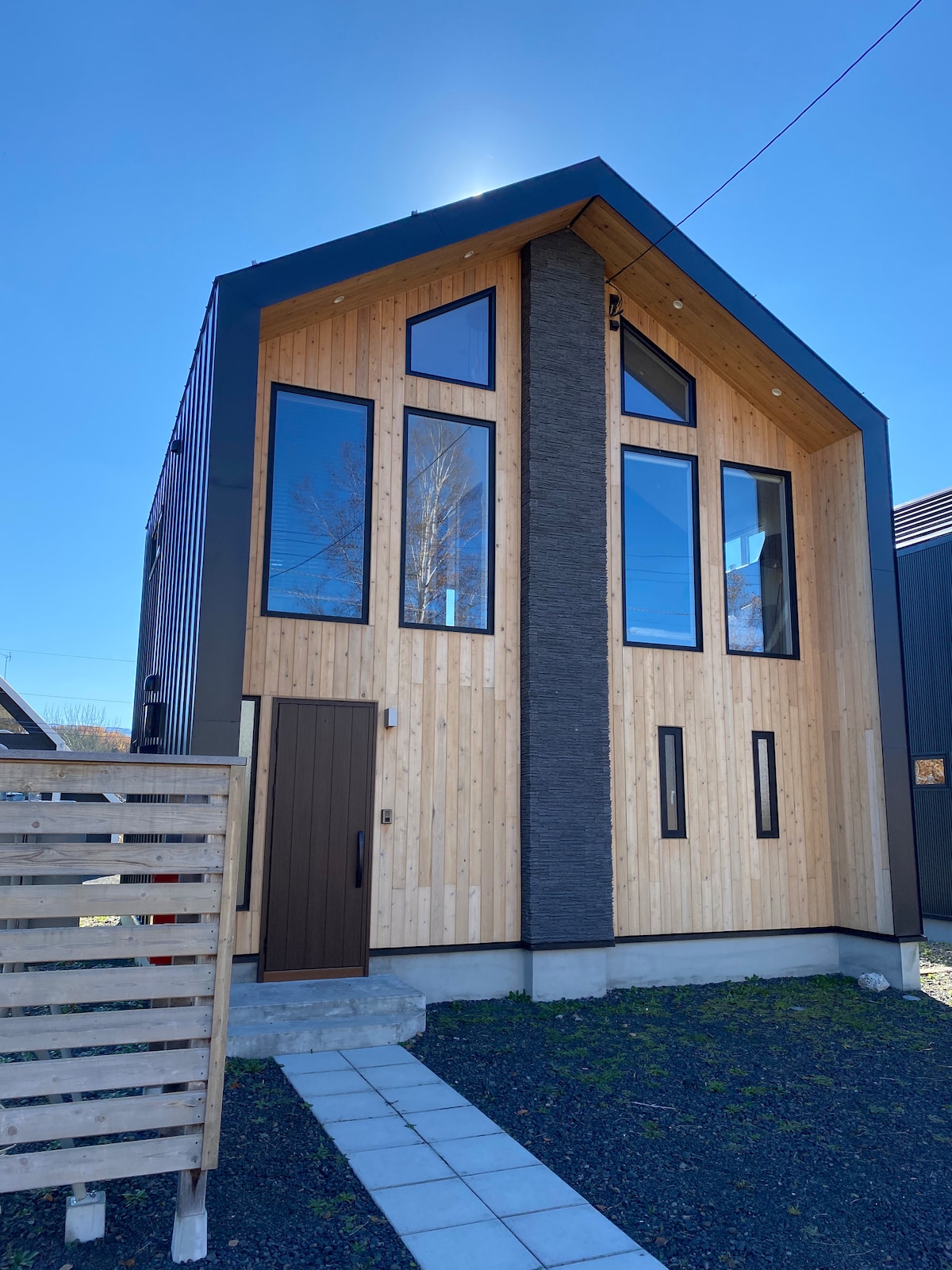 A modern home exterior is presented, featuring a combination of wood and stone elements. Large, tall windows allow natural light to enter, while a sleek front door adds a minimalist touch. A clean stone pathway leads to the entrance, framed by a wooden fence.