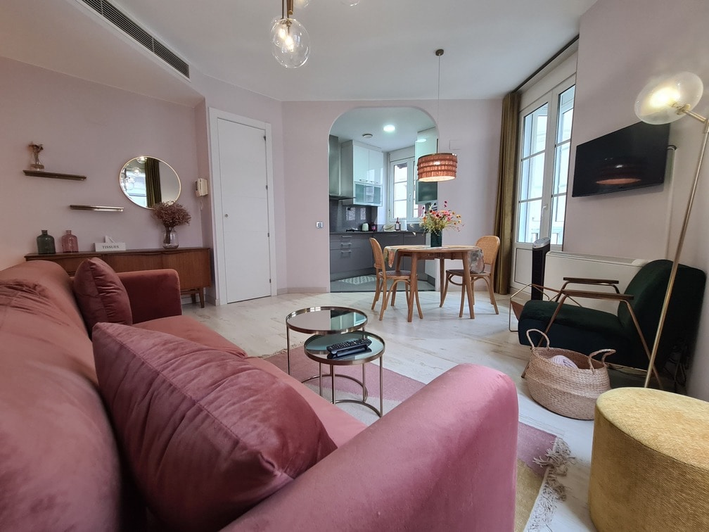 A stylish living area showcases a plush pink sofa and a round glass coffee table. A dining table is positioned nearby, accompanied by wooden chairs. Neutral walls and soft lighting create a welcoming ambiance, while a window allows natural light to enhance the space.