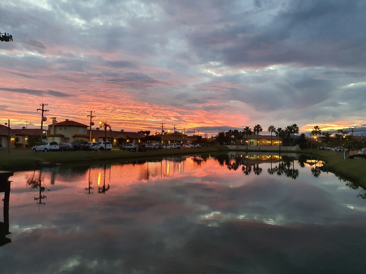 A serene lake reflects a vibrant sunset, showcasing shades of orange and purple in the sky. Surrounding buildings and palm trees are mirrored in the calm water, contributing to a peaceful ambiance. Cars are parked along the pathway, hinting at a lively community.