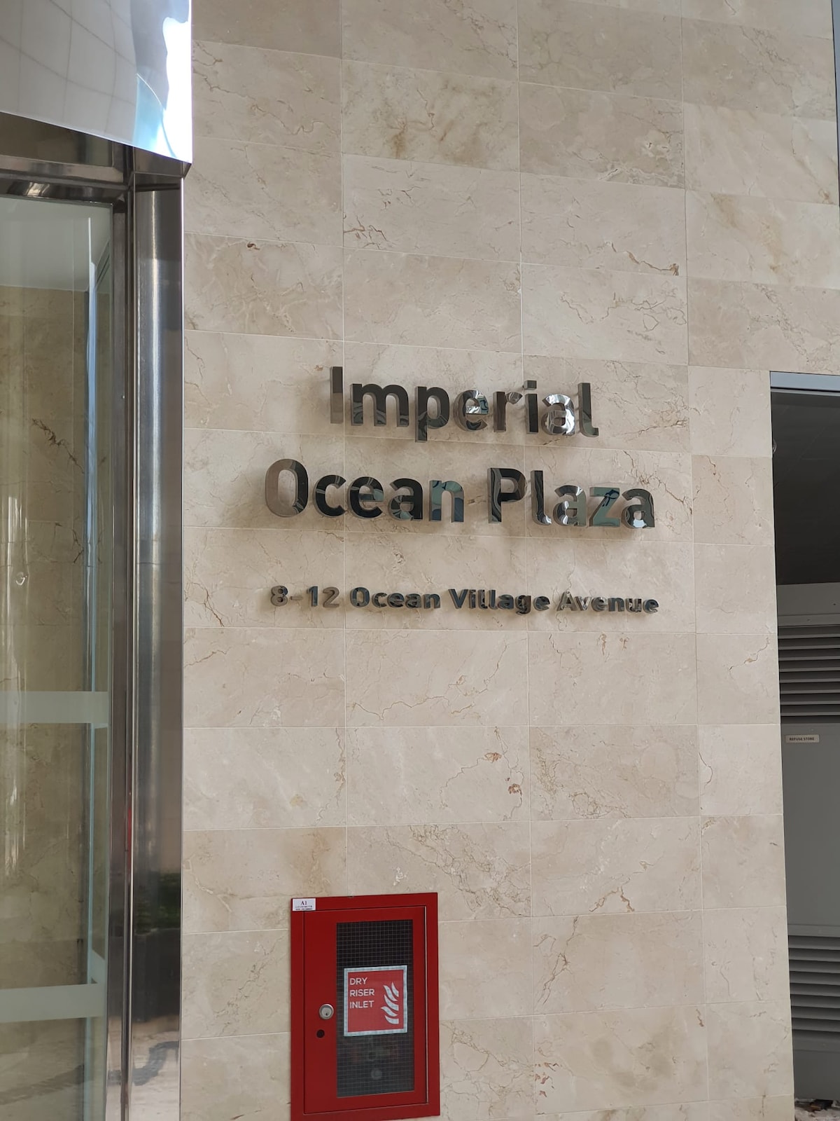 The exterior of the building features a marble wall with the text 'Imperial Ocean Plaza' prominently displayed. Below the lettering, a red fire alarm box adds a contrasting element. The entrance is highlighted by a glass door, suggesting an inviting access point.
