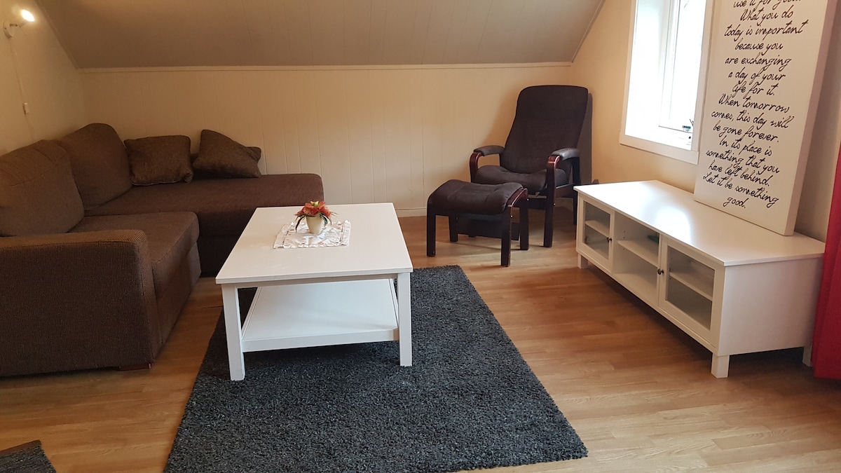 A living area features a cozy brown sectional sofa and a comfortable armchair. A white coffee table rests on a dark area rug, accompanied by a white entertainment unit. Natural light enters through a window adorned with simple curtains, creating a relaxed environment.