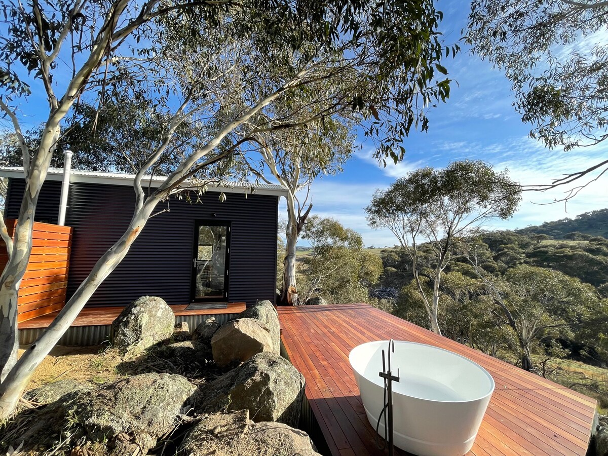 An outdoor bathing area is seen on a wooden deck, featuring a contemporary freestanding bath surrounded by large rocks and native trees. A clear blue sky complements the serene natural setting.