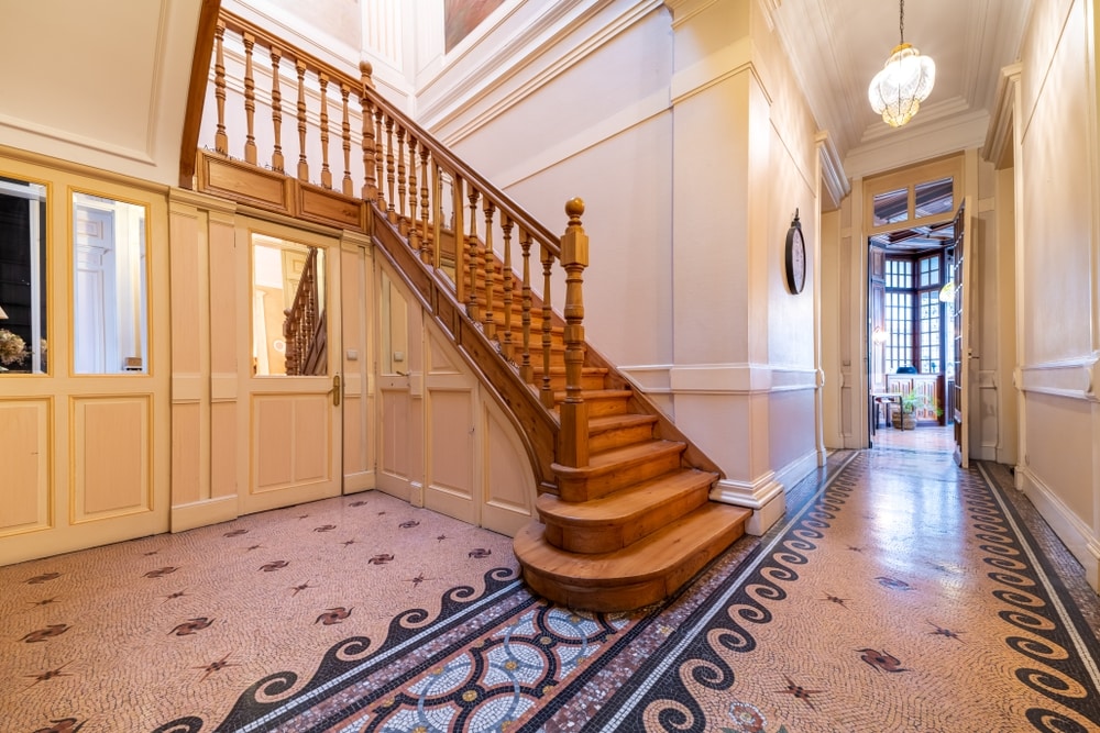 The entrance hall features a grand wooden staircase with a polished banister, leading to the upper levels. Mosaic tile flooring is displayed, accompanied by neutral walls that enhance the spacious feel of the area. Natural light is introduced through a nearby large window.