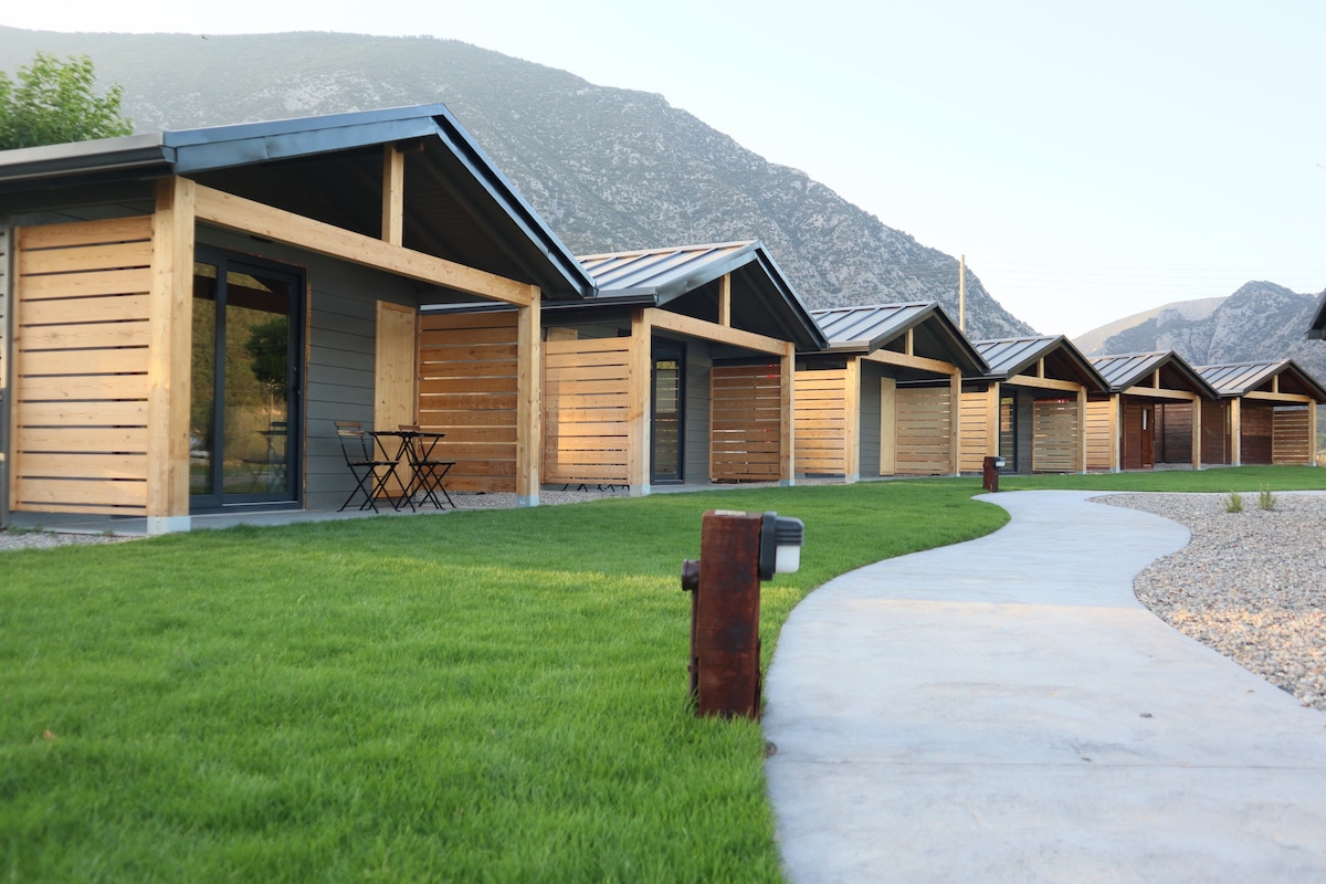 A row of wooden bungalows is situated against a mountainous backdrop. Each bungalow features large glass doors that open to a grassy area. A smooth concrete path meanders through the landscaped grounds, providing access to the bungalows.