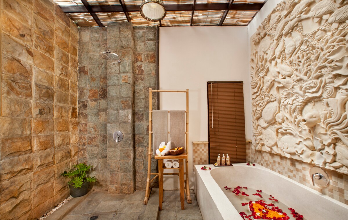 A serene bathroom features a spacious bathtub adorned with flower petals, surrounded by natural stone walls. A carved stone panel adds visual interest, while a rainfall shower is situated nearby. A bamboo chair and neatly arranged toiletries complete the calming atmosphere.