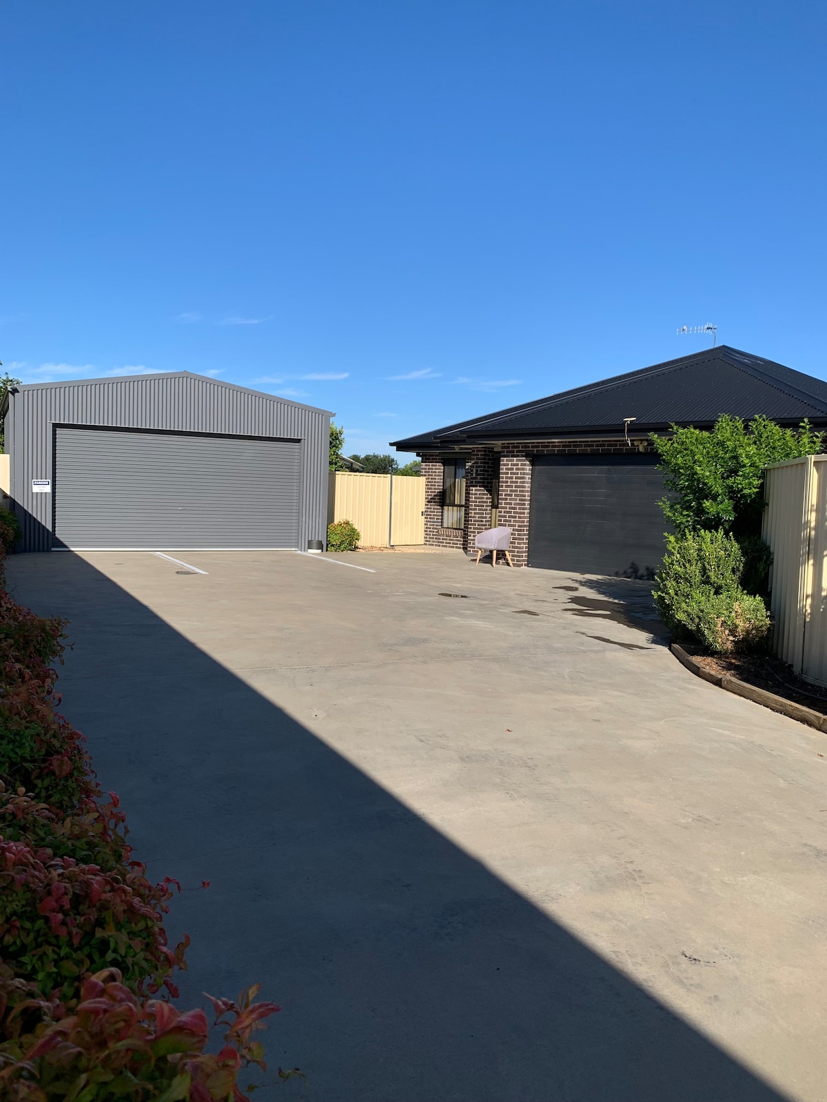 A driveway leads to a residential property, featuring a detached garage and a single-story house with a dark roof. Lush green shrubs line the perimeter, while a shaded area provides a cozy seating option. Clear blue skies are visible overhead.