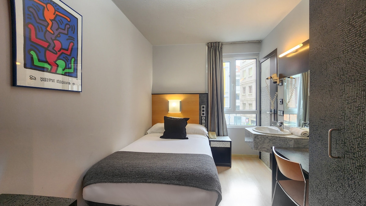 A modern single room is presented, featuring a spacious bed with a dark throw pillow. Natural light enters through a large window, and a contemporary artwork adorns the wall. The room includes a compact desk and an attached private bathroom with minimalist design elements.