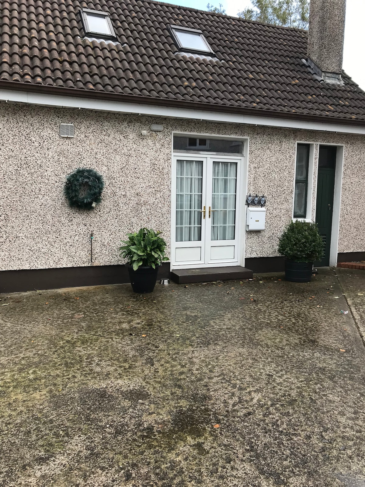 The entrance features a pair of glass-framed doors flanked by potted shrubs. A textured exterior wall and a small decorative wreath add subtle charm, while a clear path leads to the inviting entry.