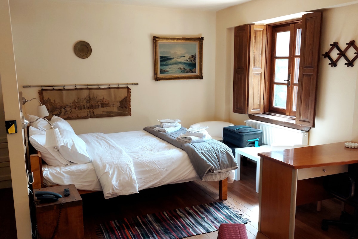 A cozy sleeping area features a spacious bed with white linens, accented by a soft gray throw. Natural light fills the room through wooden windows, while art and textiles add warmth. A small desk and suitcase are also present, contributing to the welcoming space.