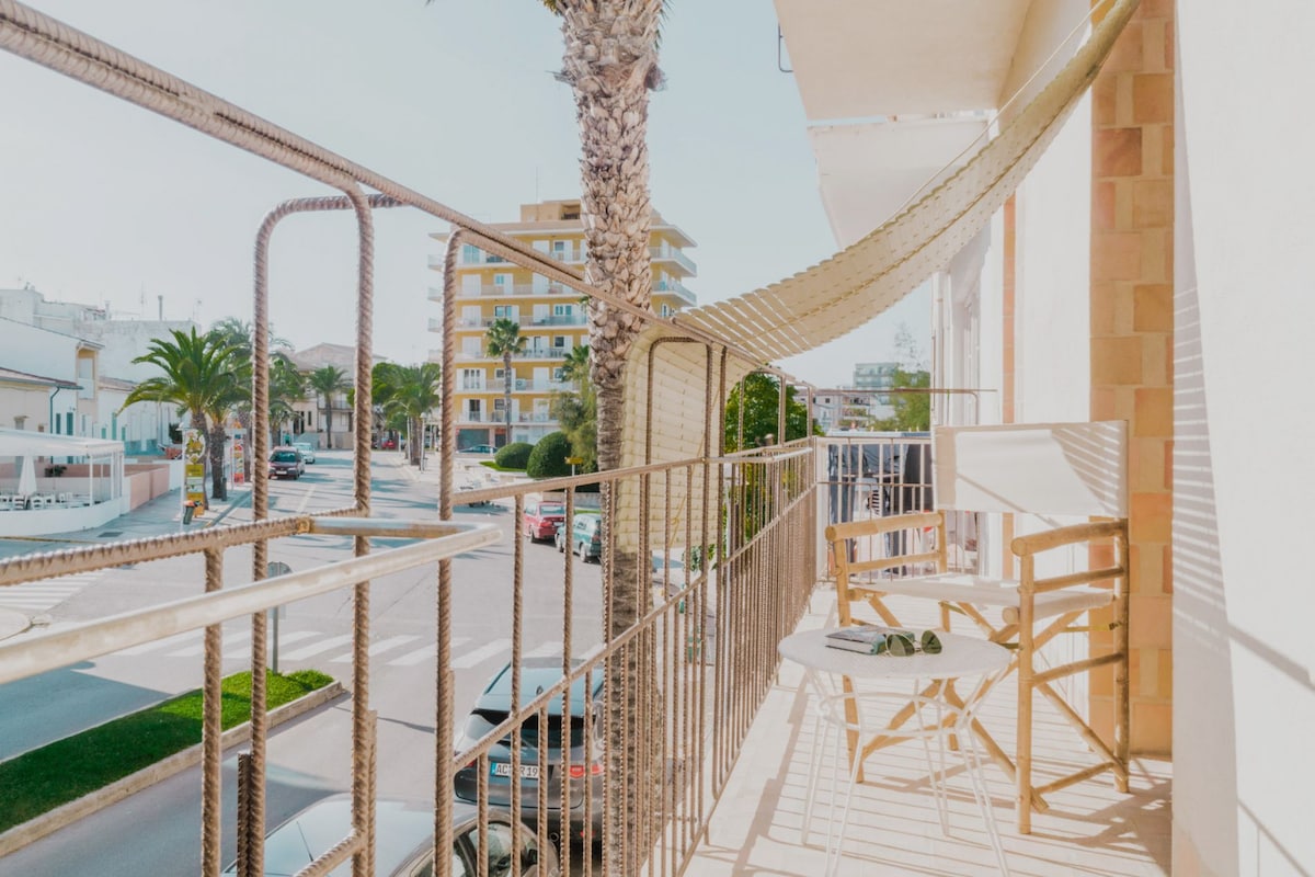 A balcony is depicted with a light-colored railing, featuring two wooden chairs and a small table. Sunlight casts soft shadows on the floor, while palm trees and buildings are visible in the background, providing a glimpse of the surrounding area.