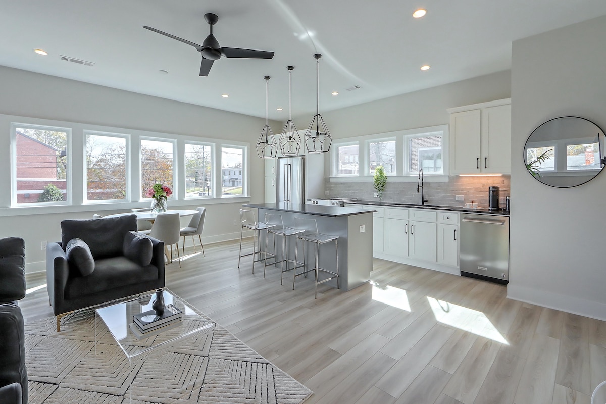 A spacious open-concept living area showcases a modern kitchen with stainless steel appliances and a large island, providing ample seating. Natural light fills the room through multiple large windows, enhancing the airy feel. A comfortable seating area with a dark sofa is positioned nearby.