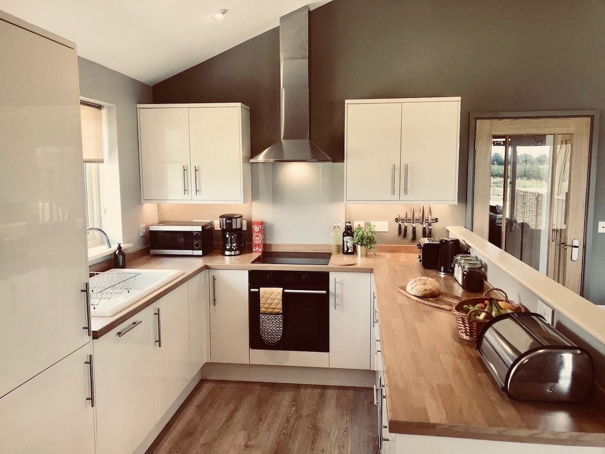 A modern kitchen is presented with sleek white cabinetry and a spacious countertop. Equipped with essential appliances, including a stove and microwave, the area is illuminated by natural light. A wooden table is set with baked goods, enhancing the inviting atmosphere.