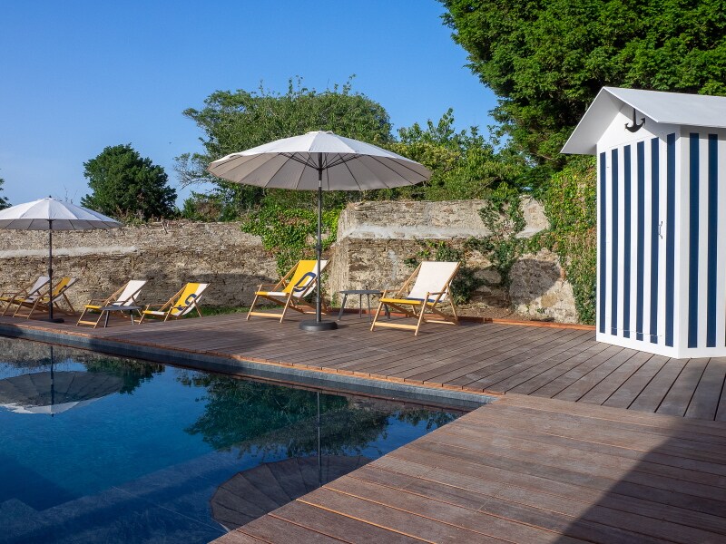 A serene swimming pool area features wooden decking surrounded by lounge chairs under large umbrellas. A storage shed with blue and white stripes is positioned nearby, complemented by lush greenery and a stone wall, creating a calm outdoor environment.