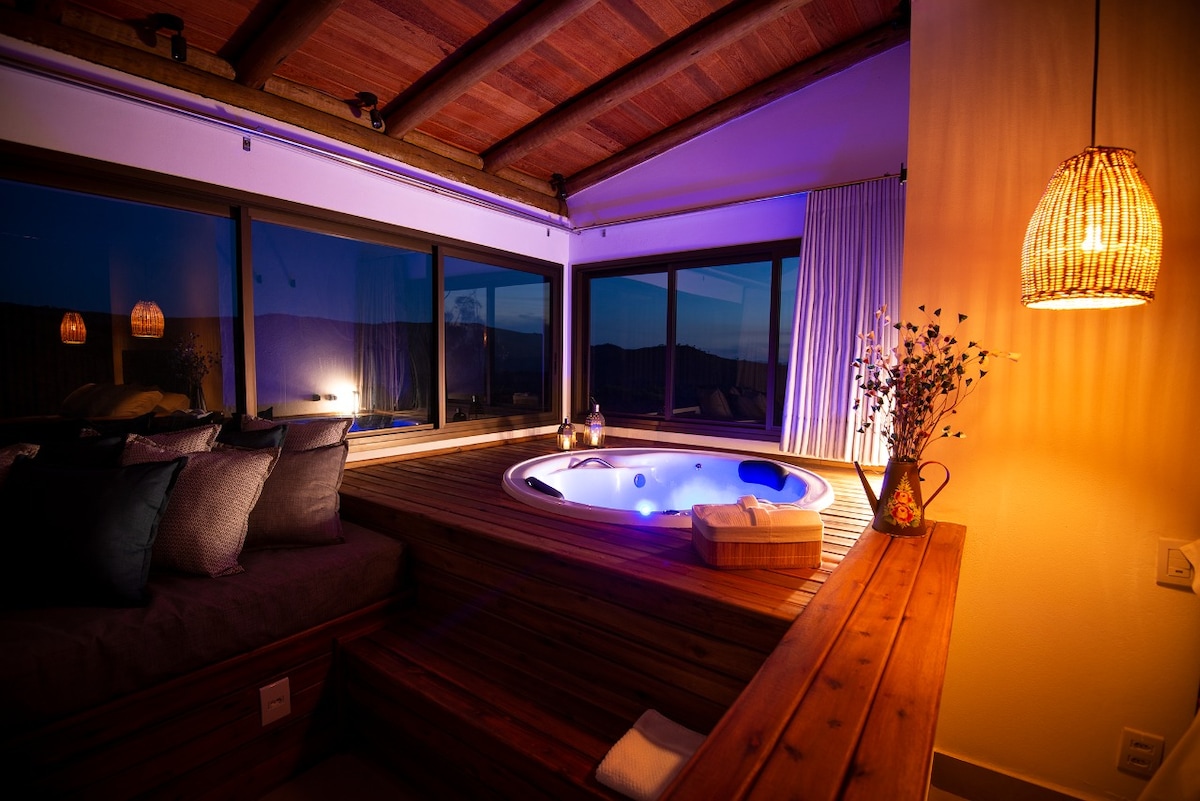 A serene space features a hot tub surrounded by wooden decking, with soft, ambient lighting creating a calming atmosphere. Large windows provide expansive views of the outdoor landscape, while plush cushions are arranged nearby for relaxation.