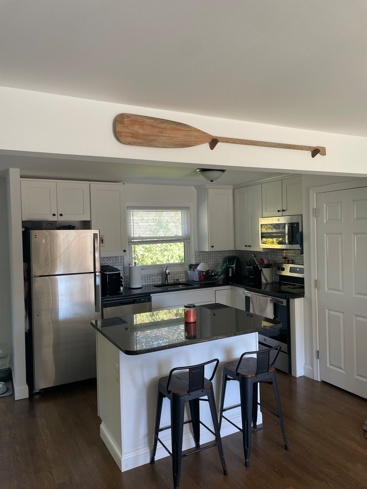 A modern kitchen features stainless steel appliances, including a refrigerator and oven. The black countertop offers a workspace, with two metal stools positioned nearby. Light enters through a window, while a wooden paddle is displayed above the cabinetry, adding a subtle decorative touch.
