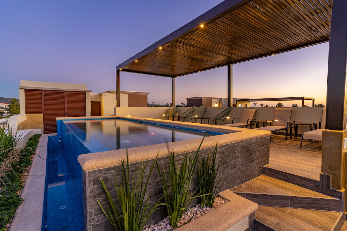 An outdoor terrace features a tranquil pool under a shaded structure. Comfortable lounge chairs are positioned around the pool, accompanied by landscaped greenery. Soft, ambient lighting enhances the serene atmosphere as dusk approaches, offering panoramic views of the surrounding area.