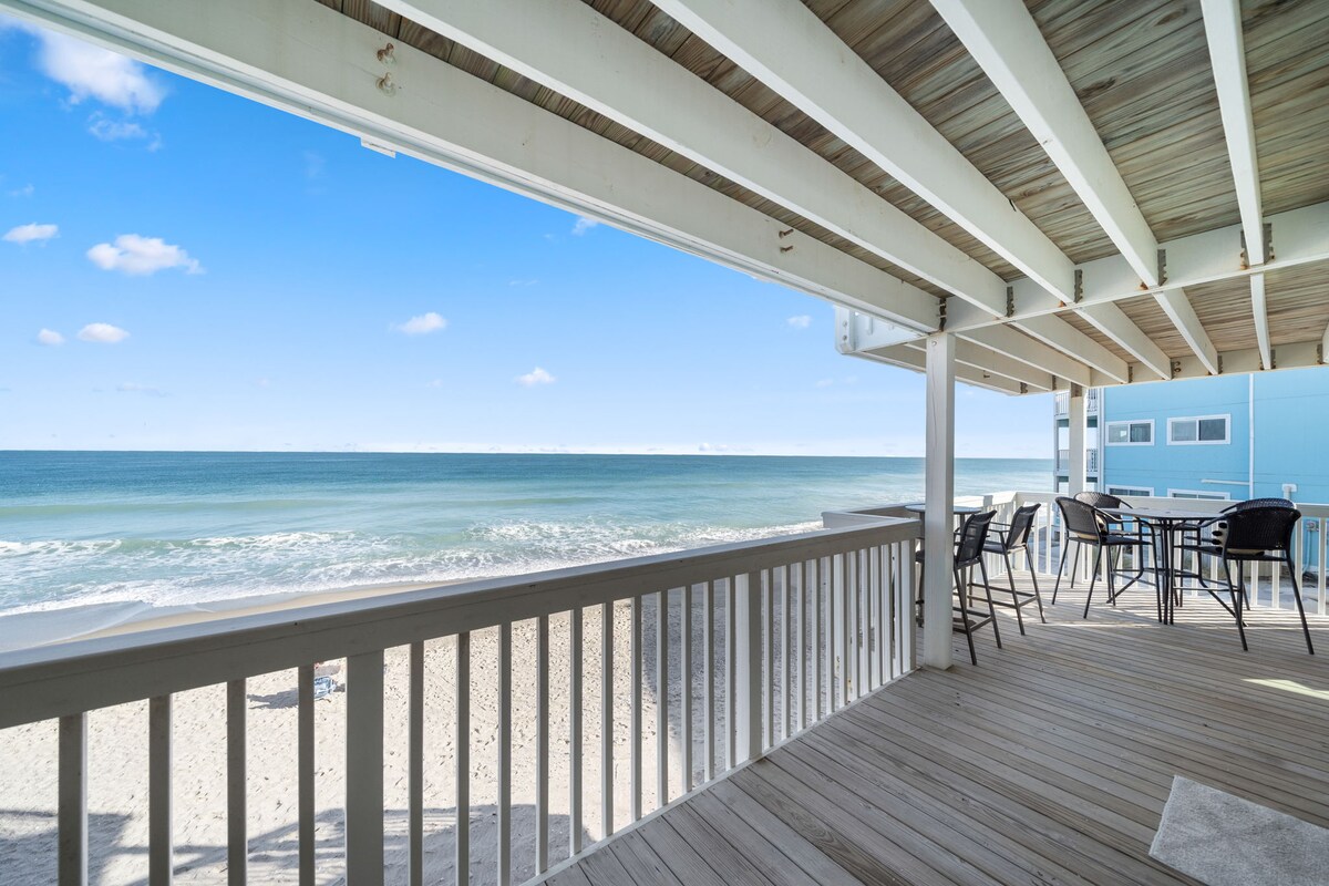 The oceanfront balcony presents expansive views of the sandy beach and waves. A wooden railing encircles the balcony, complemented by a seating area including high-top chairs and a table. Bright blue skies and a few fluffy clouds add to the serene coastal atmosphere.