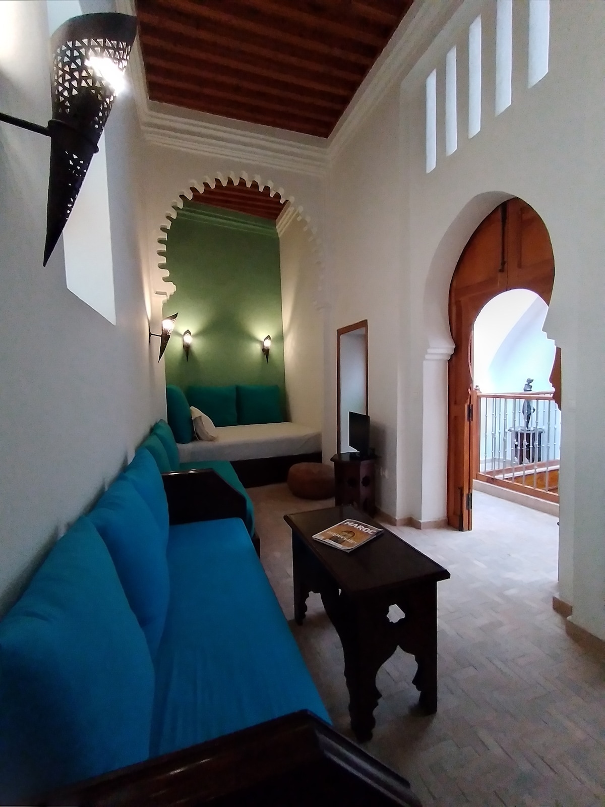 A seating area features turquoise sofas against a backdrop of whitewashed walls and intricately designed arches. Light fixtures add warmth, while a wooden table with a magazine provides functionality. An inviting sleeping area is visible through the open doorway, enhancing the sense of space.
