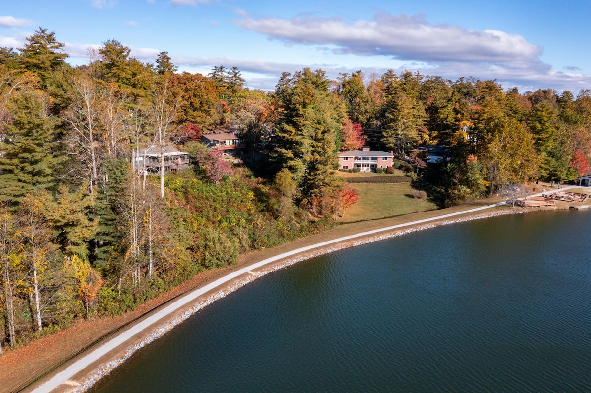 A serene view showcases the cabin nestled among lush trees with vibrant fall foliage. The tranquil water mirrors the colorful landscape, complemented by a scenic walking path along the water's edge. The combination of nature and architecture creates a peaceful ambiance.