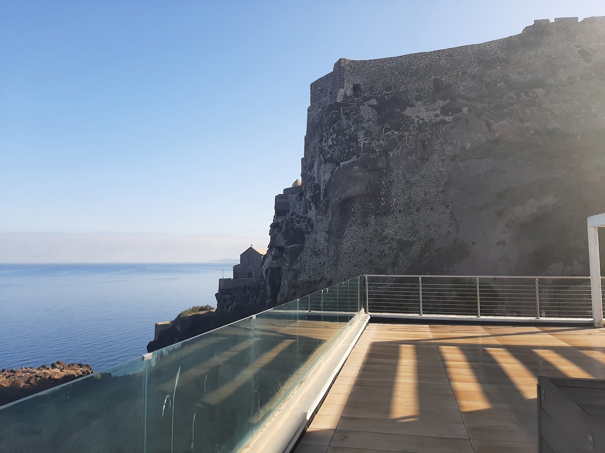 A panoramic view reveals a serene sea meeting a clear sky, with a fortress-like structure perched on a rocky cliff. The terrace glass railing provides unobstructed sightlines, while soft shadows are cast across the wooden decking from the surrounding architecture.