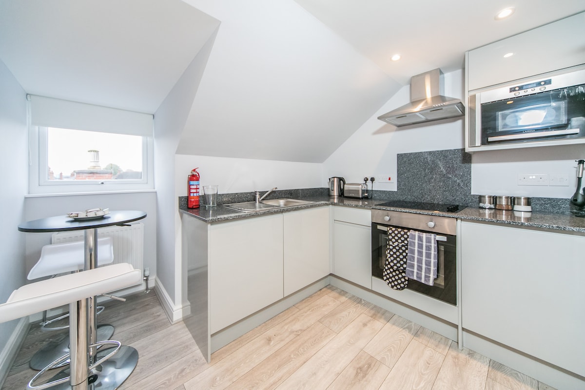 A modern kitchen features sleek cabinetry and a gray countertop. A stovetop and microwave are integrated into the design, while a kettle, utensils, and other essentials are neatly arranged. A small round dining table with two barstools is situated in one corner, enhancing the space's functionality.