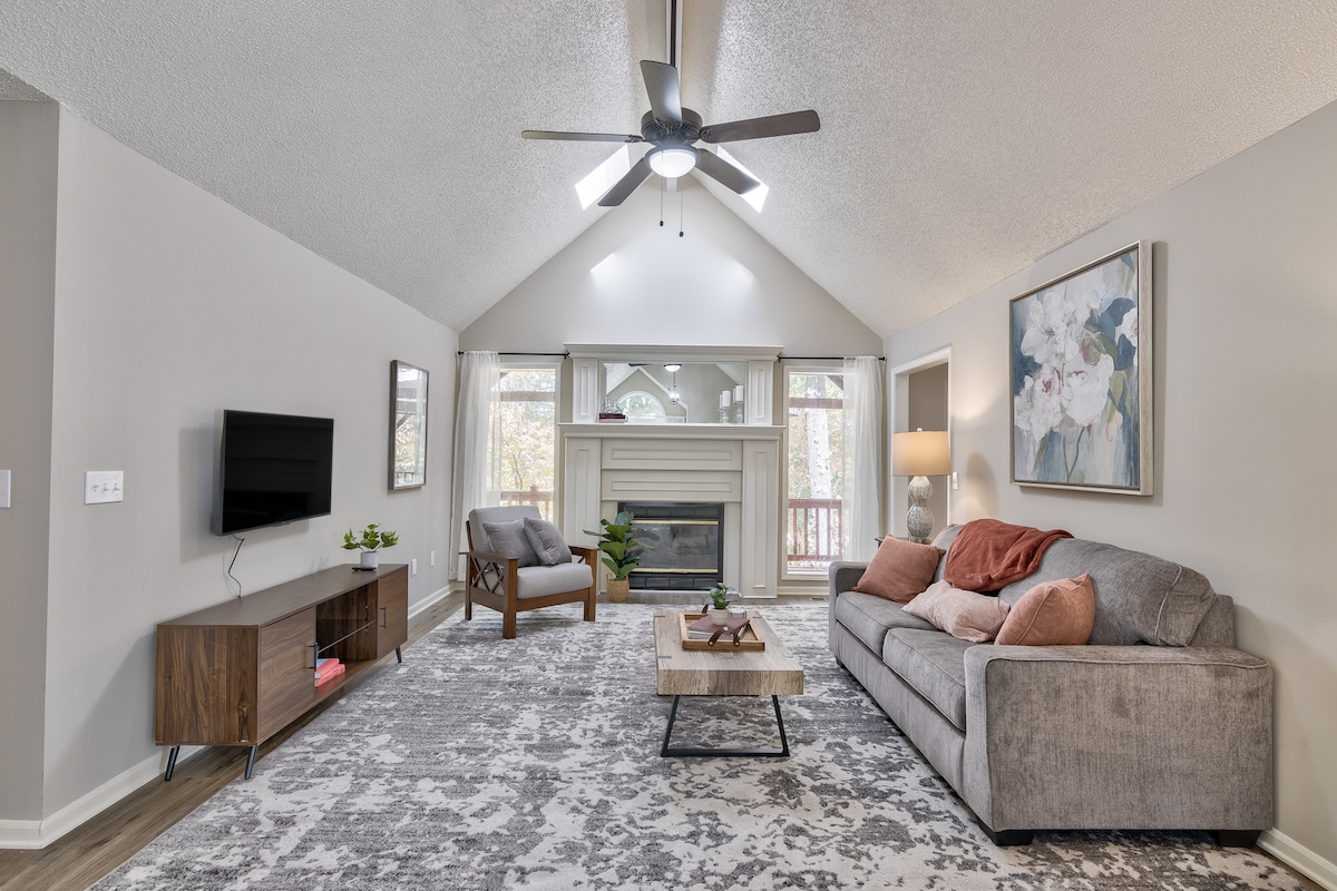 A spacious living room is highlighted by a large light fixture and a ceiling fan. A comfortable gray sofa and a wooden armchair are situated near a stylish coffee table. A mounted TV and a modern fireplace add to the inviting space, with large windows overlooking the deck.