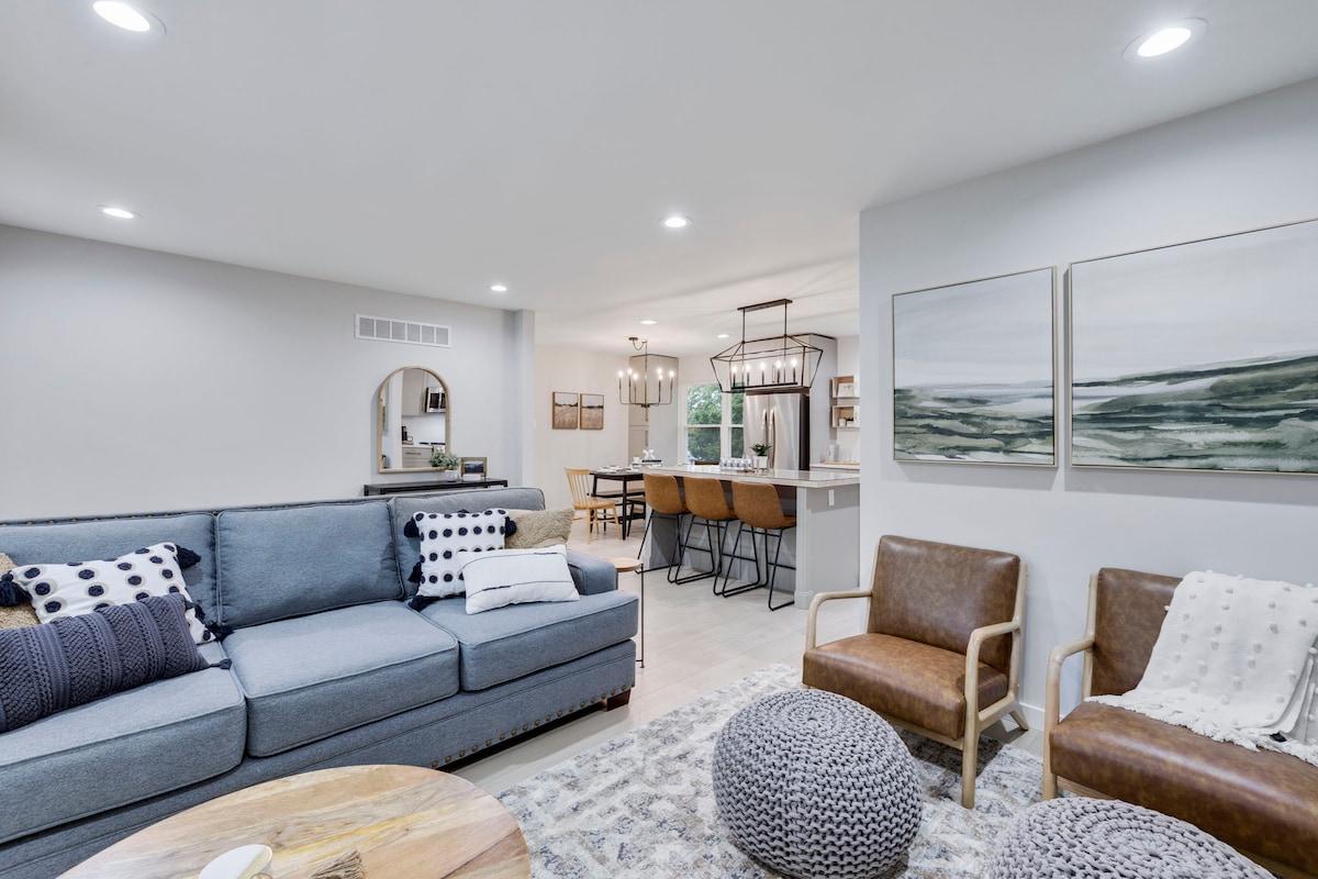 A modern living area features a blue sectional sofa adorned with patterned pillows. Two brown accent chairs are positioned nearby, complemented by round poufs. A dining area is visible in the background, illuminated by overhead lighting, with a large table and stylish chairs.
