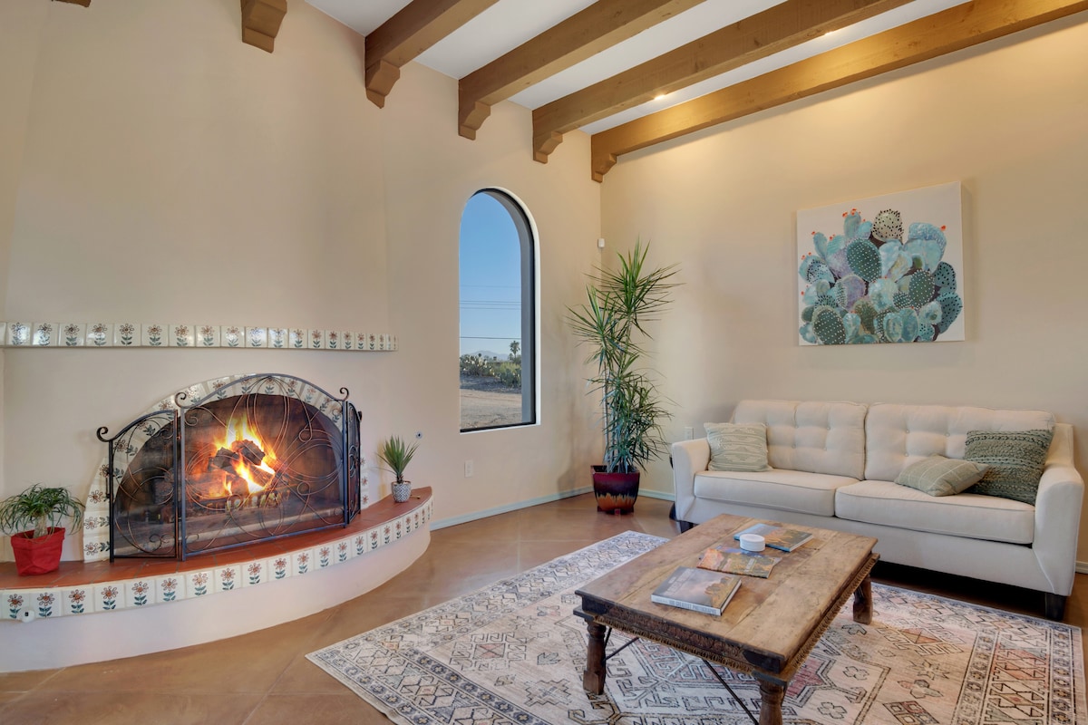 A main living area features a cozy beehive gas fireplace with decorative tile work and an arched window allowing natural light. A light-colored couch is positioned beside a rustic wooden coffee table, enhancing the inviting atmosphere. Potted plants add a touch of greenery.