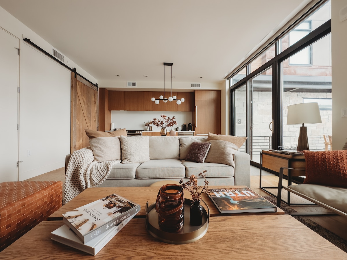 A spacious living area is featured, centered around a light-colored sofa adorned with soft cushions. A wooden coffee table holds decorative items, including magazines and a tray. Large windows provide ample natural light, framing a view of the modern kitchen in the background.