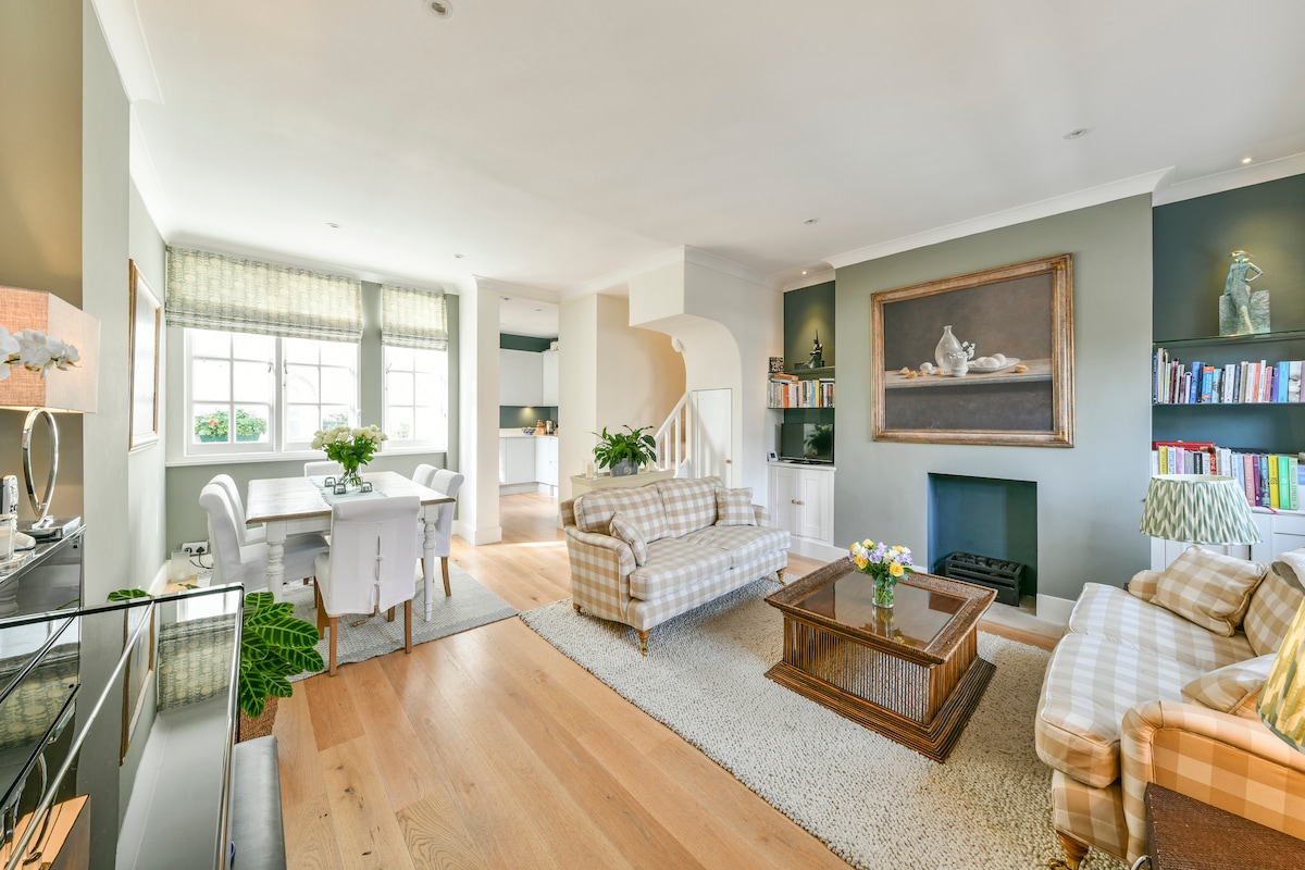 A spacious living area features a comfortable seating arrangement with patterned sofas and a coffee table at its center. Natural light filters through large windows, illuminating a dining table set for four. A bookshelf with a variety of books is positioned against the wall near a cozy fireplace.