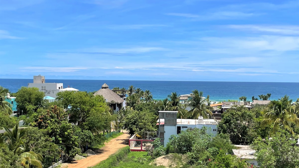 A panoramic view showcases the vibrant landscape of Zicatela Beach, featuring lush greenery and a distant ocean. The scene includes a thatched-roof palapa, tropical palm trees, and distant structures under a clear blue sky.
